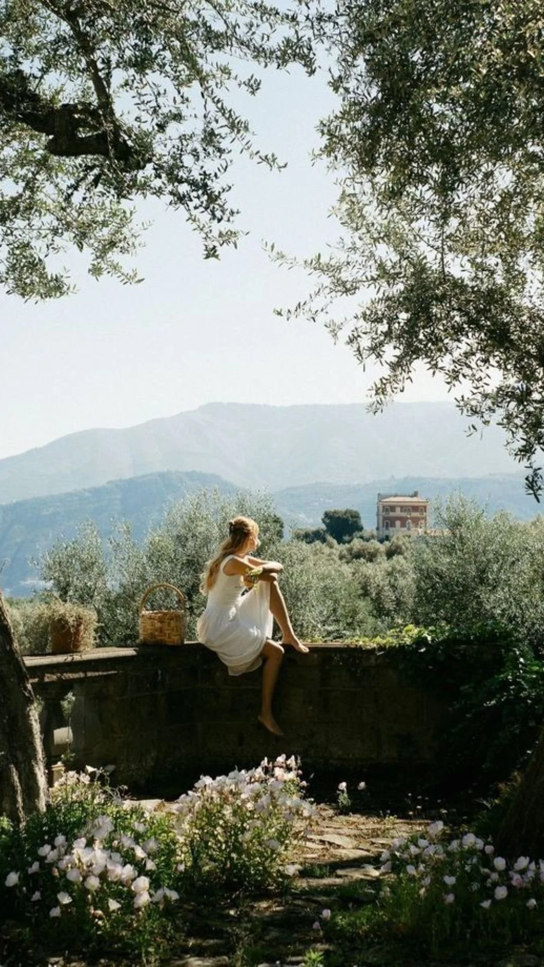 A woman in a white dress sitting on a stone wall, surrounded by greenery and flowers, with a basket nearby. Mountains can be seen in the distance under a clear sky.
