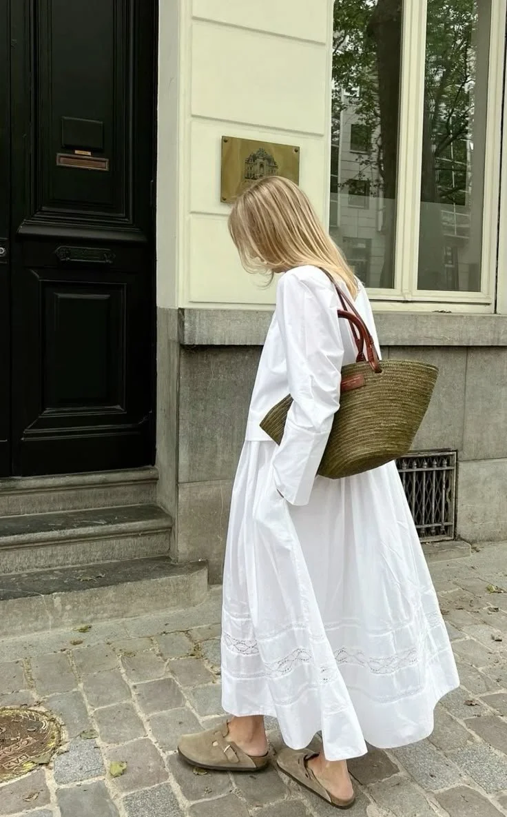 A woman standing on a cobblestone sidewalk outside a beige building with large windows, wearing a long white dress, tan slip-on shoes, and carrying a large straw bag.