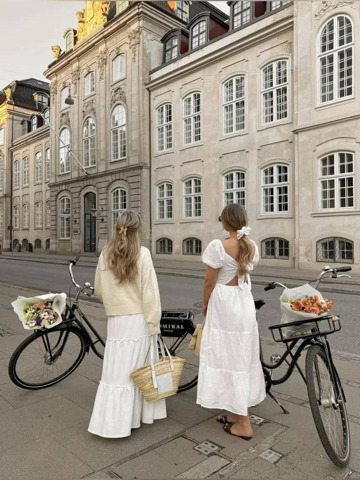 Two women dressed in white dresses stand on a city sidewalk beside two bicycles, each with flowers in the baskets. They look at a historic, ornate building with large windows and decorative architectural details.