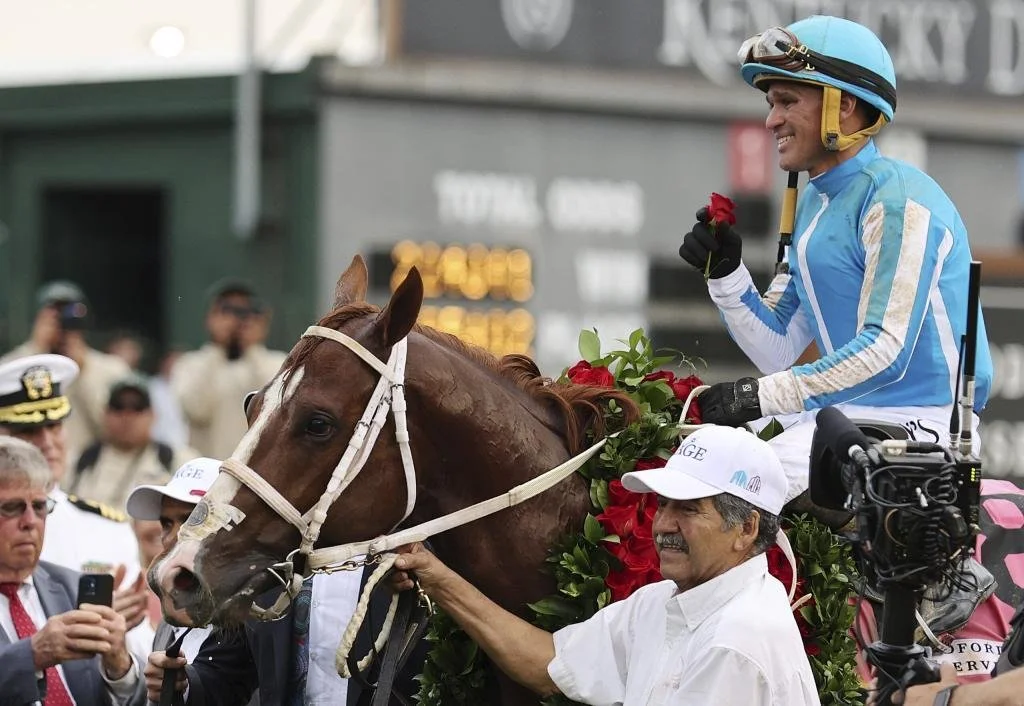 Kentucky derby winner -Jockey in blue silks on a brown horse holding a red flower, surrounded by people including a man with a white cap and others in suits, at a racing event.