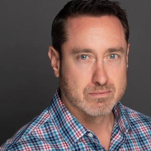 Close-up portrait of a middle-aged man with blue eyes, short dark brown hair, light beard, wearing a blue and red checkered shirt, against a dark gray background.