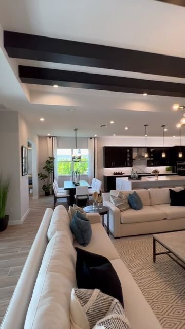 Open living room and kitchen with ceiling beams, large window, and modern furniture, including a beige sectional sofa with pillows, a dining table with chairs, and a kitchen with dark cabinets and pendant lights.