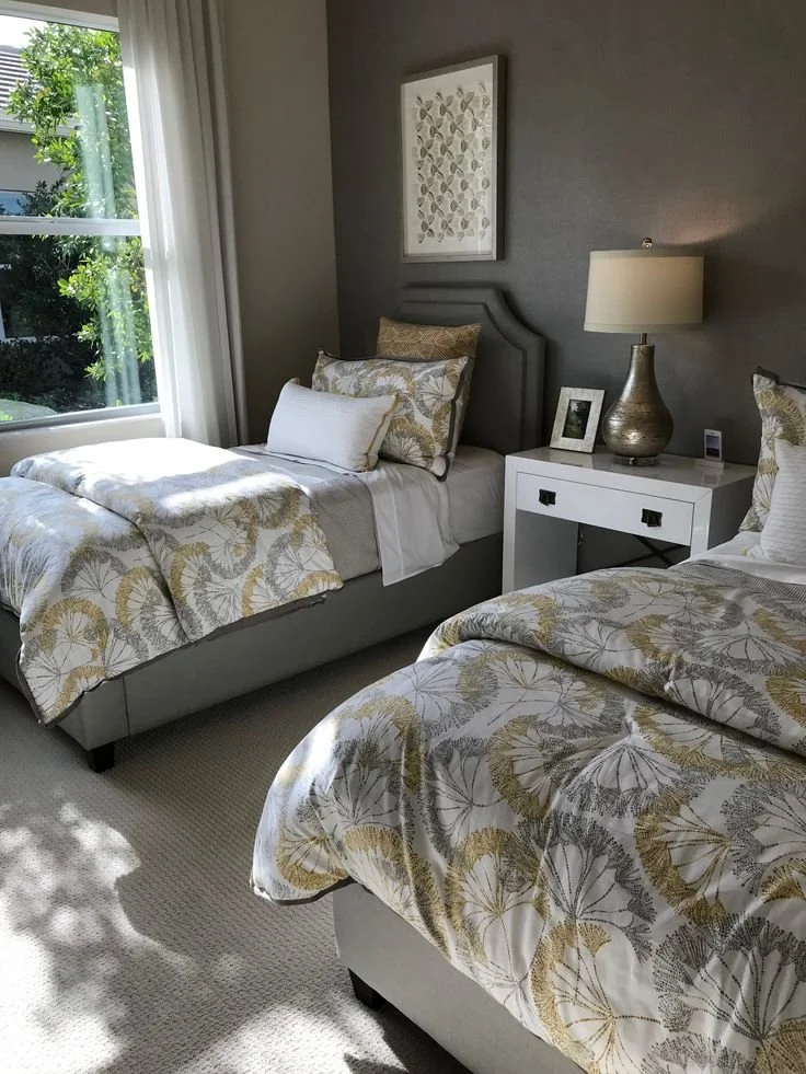 Two twin beds with patterned bedding and pillows in a bedroom near a window with white curtains. A nightstand with a lamp, framed photo, and small electronic device sits between the beds. There is wall art above the headboard of one bed.