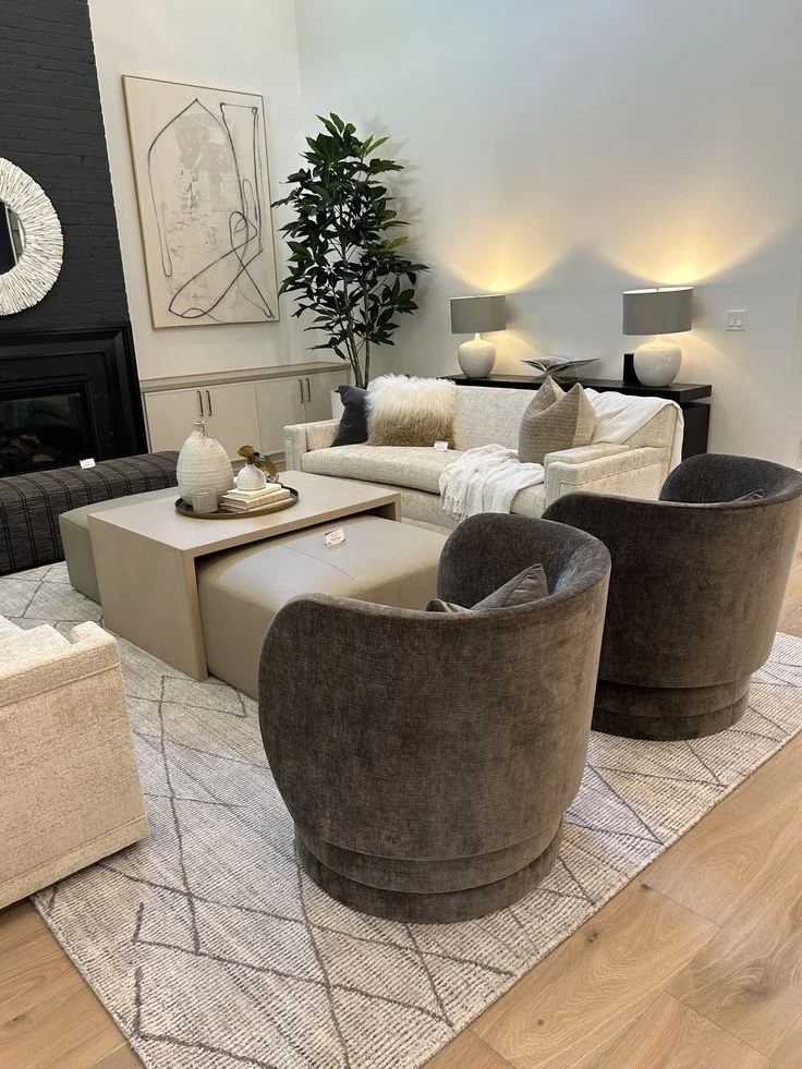 Modern living room with beige and gray furniture, a white sofa with throw pillows, a black coffee table, and two gray armchairs. There are decorative lamps on side tables, a large green indoor plant, abstract wall art, and a patterned area rug over wood flooring.