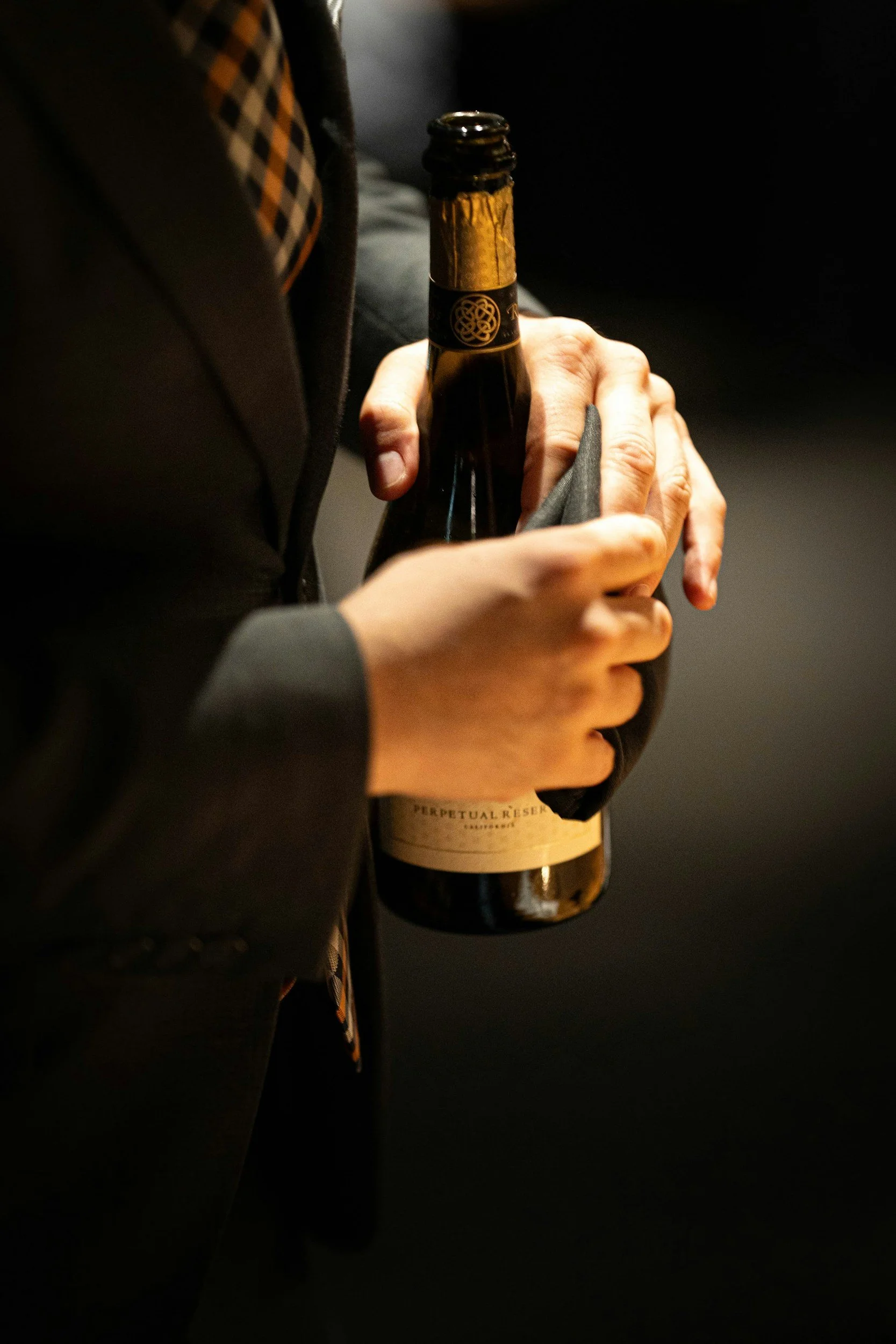 Person in a suit holding a dark-colored wine or champagne bottle in their hands.