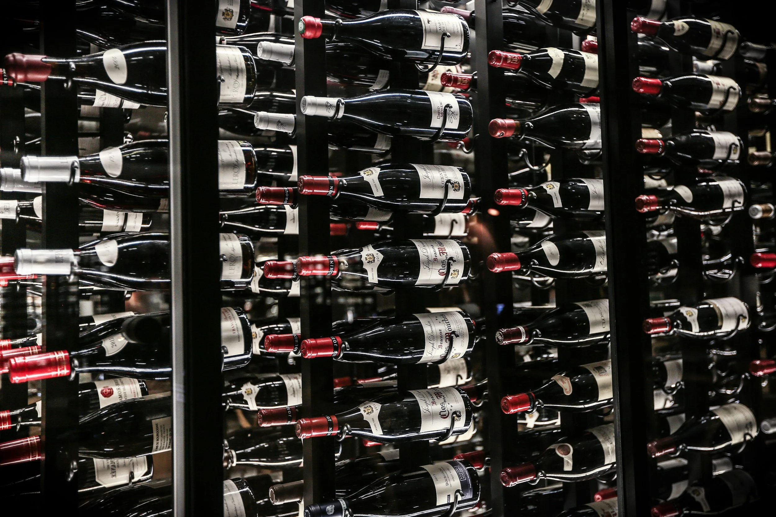 A wine cellar with multiple rows of horizontal wine bottles stored on black racks, with red-tipped bottle caps.