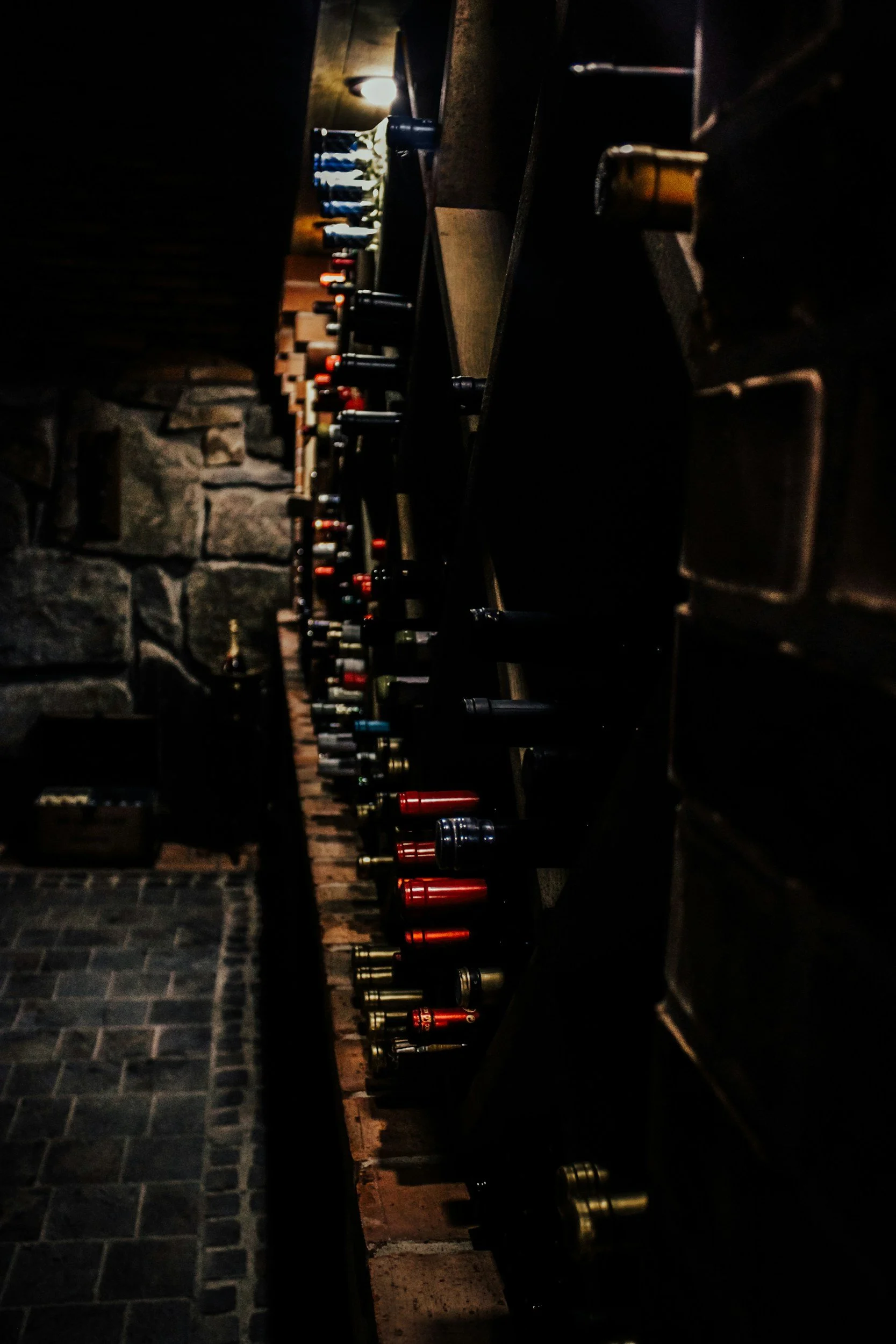 A dark room with a wine rack holding multiple bottles of wine, with a stone wall and a dim light visible in the background.