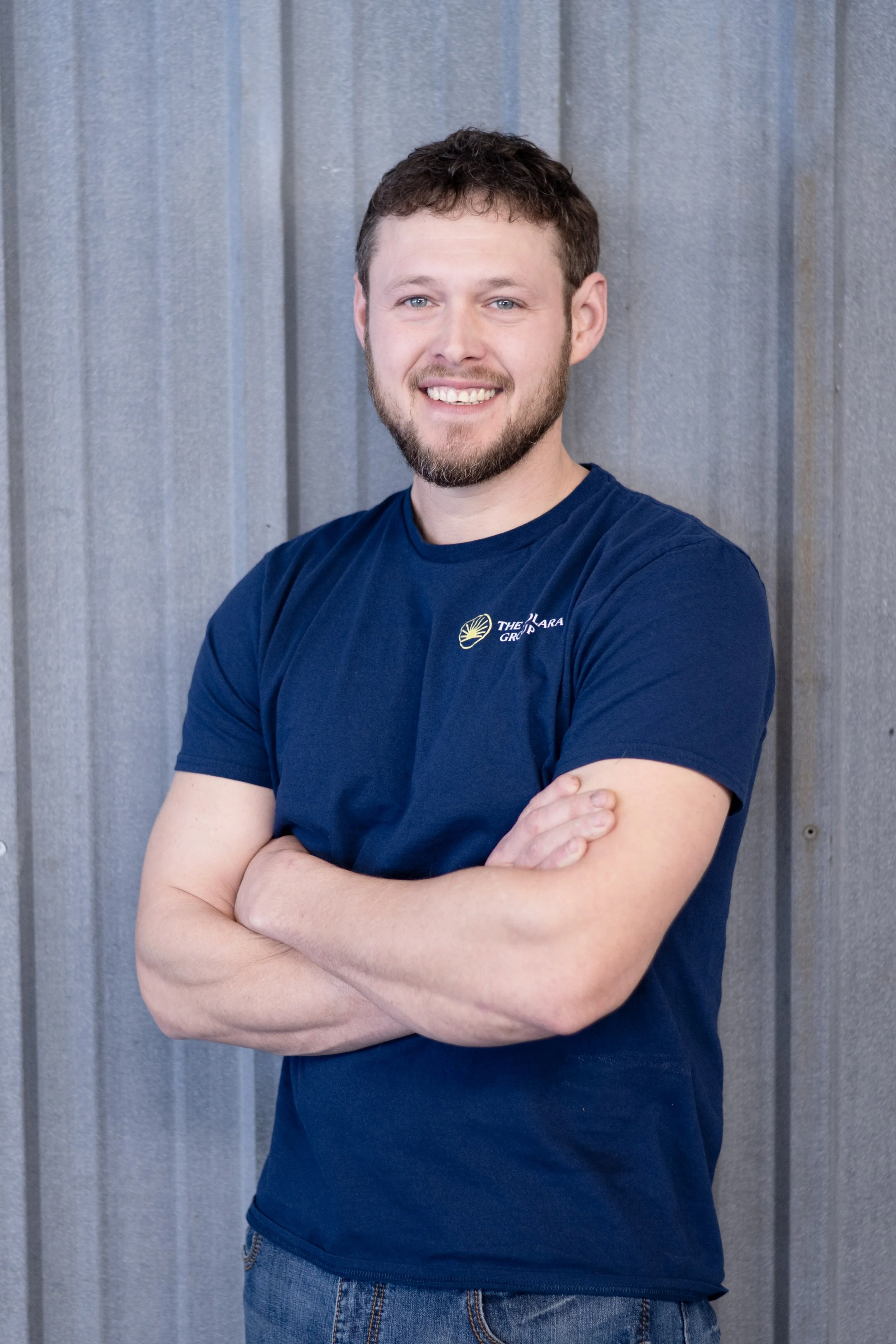 Smiling young man with a beard and mustache, wearing a navy blue t-shirt with a logo, standing with arms crossed in front of a metal wall.