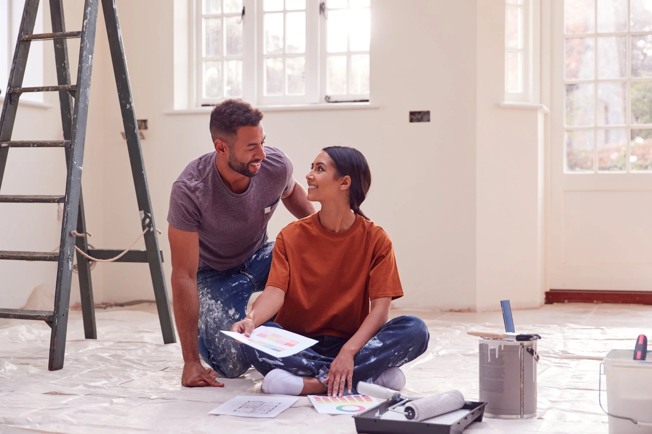 A couple working together on home renovation inside a bright room with large windows, sitting on the floor surrounded by painting supplies, including a paint roller and color swatch, with a ladder nearby.