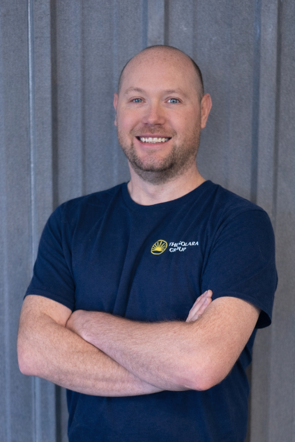 A smiling man with a shaved head and beard, wearing a navy blue T-shirt with a yellow and white logo, standing against a gray wooden wall with arms crossed.