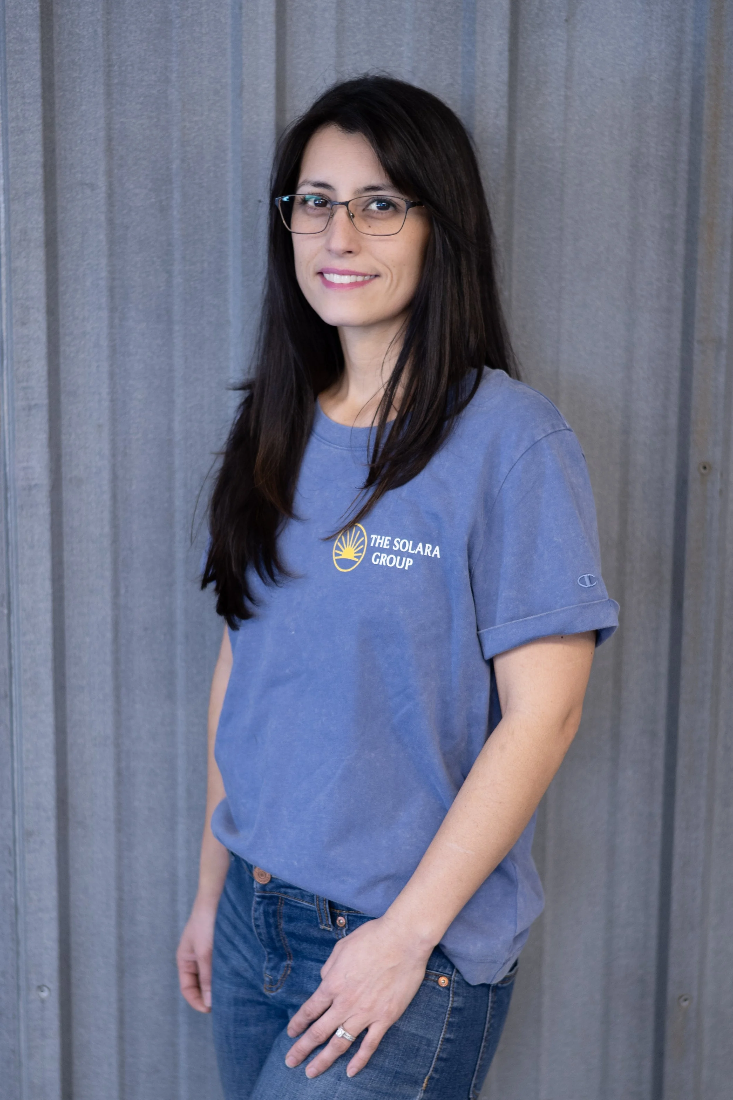 A woman with long dark hair and glasses wearing a blue T-shirt with 'The Solara Group' logo, standing in front of a gray corrugated metal wall.