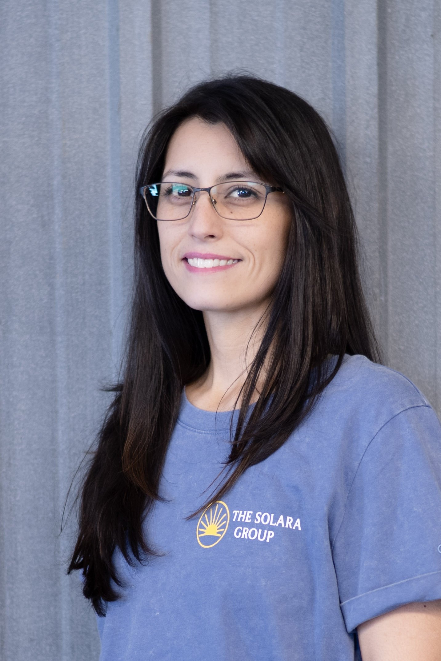 A professional woman with shoulder-length dark hair and glasses, wearing a dark blazer, standing outdoors with arms crossed in front of a modern office building.