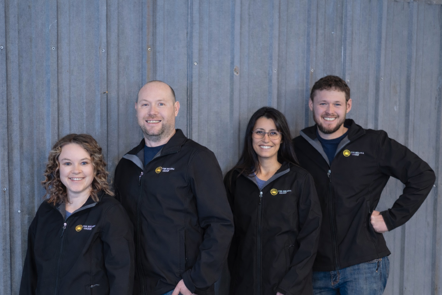 Group of four smiling individuals standing against a metal wall, wearing black jackets with a logo that reads 'THE SOLARA GROUP'.
