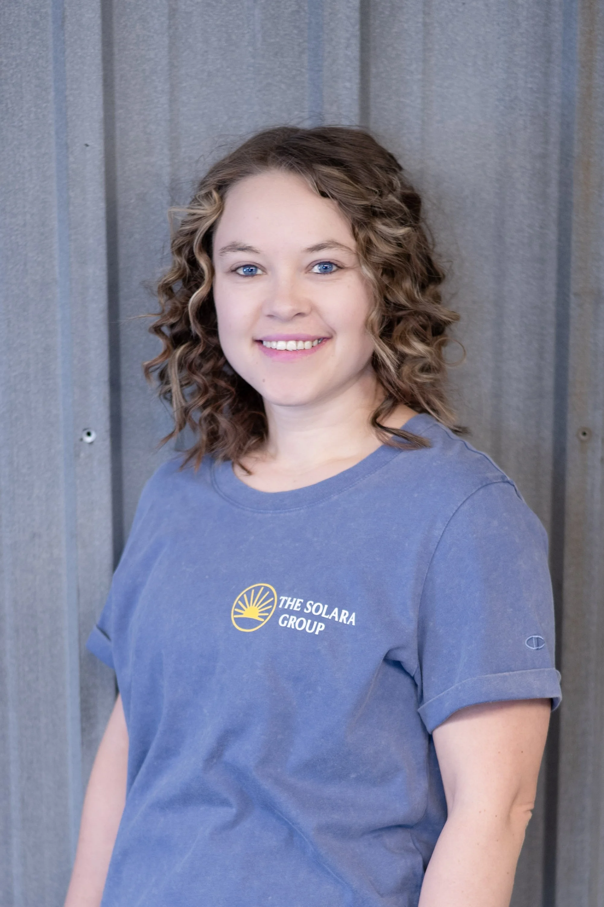 A woman with curly brown hair and blue eyes wearing a blue t-shirt with 'The Solara Group' logo, standing in front of a grey corrugated metal wall, smiling at the camera.