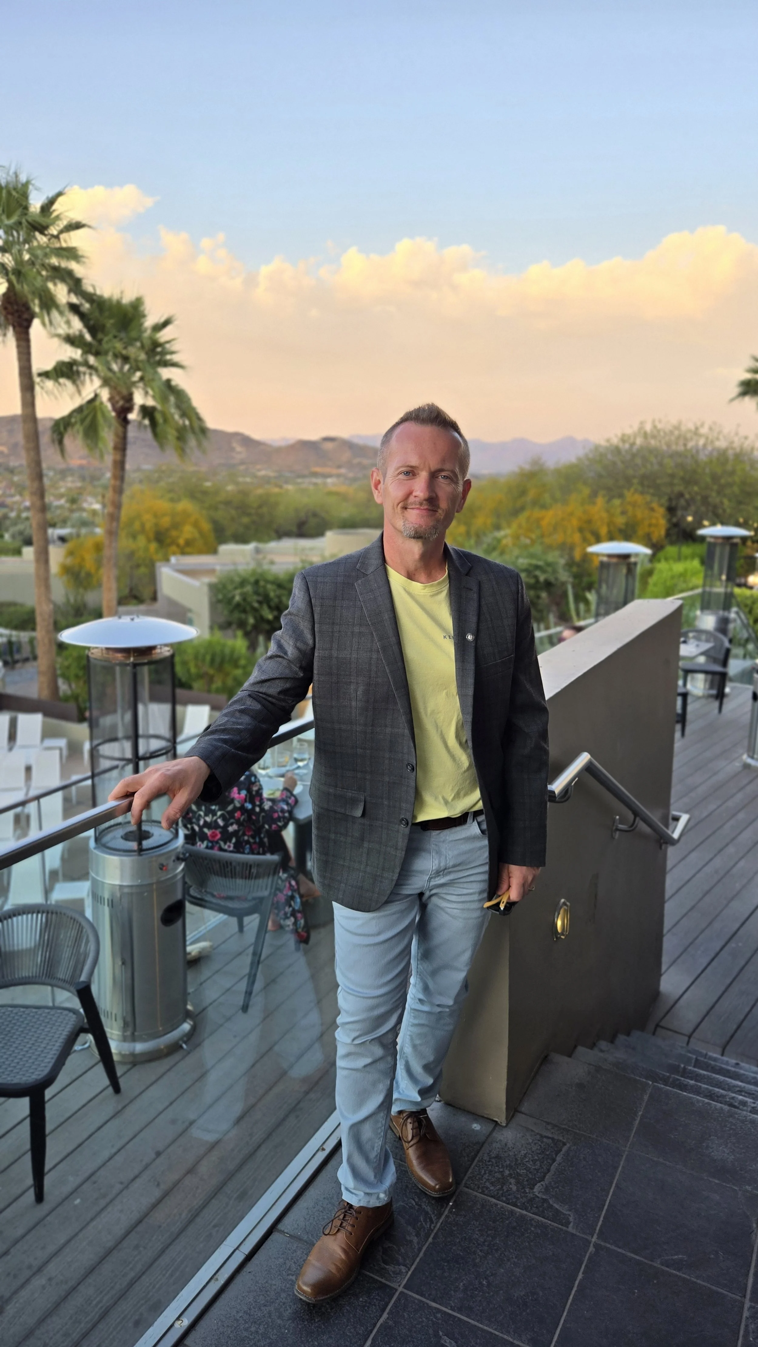 A man wearing a dark plaid blazer, yellow t-shirt, light blue jeans, and brown shoes standing on an outdoor terrace at sunset, with palm trees and mountains in the background.
