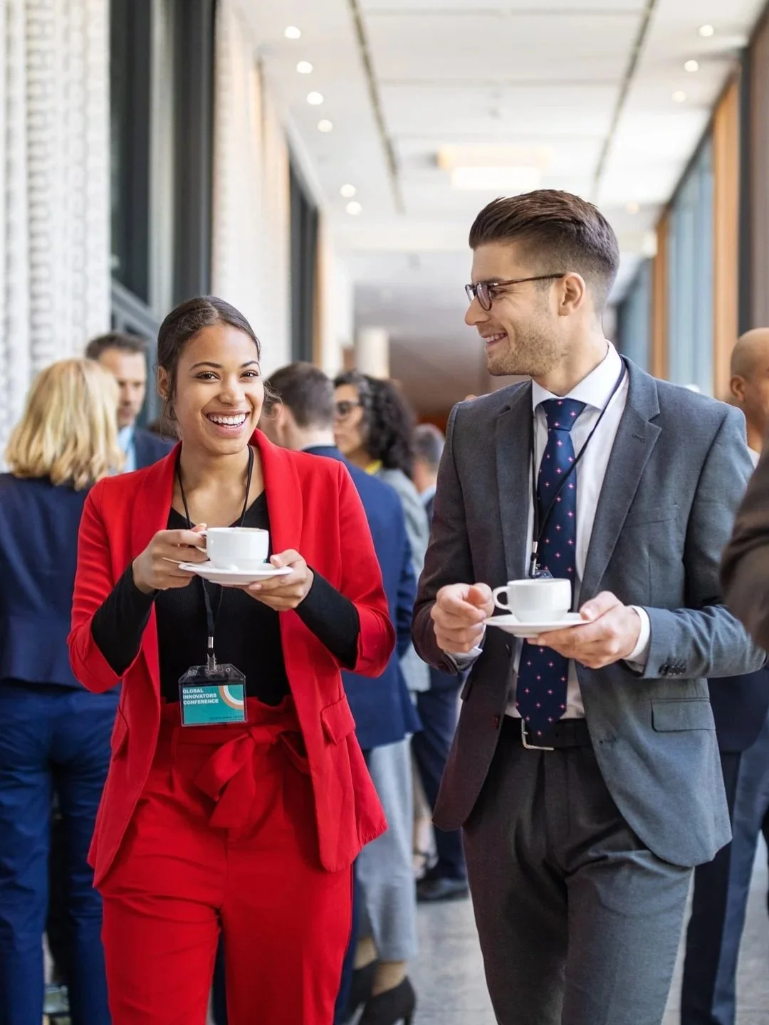 Two Network Marketing professionals having a conversation at a conference, holding cups of coffee, with conference badges, in a modern, well-lit venue.
