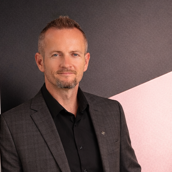 A man in a dark blazer and black shirt standing against a dark and light background.