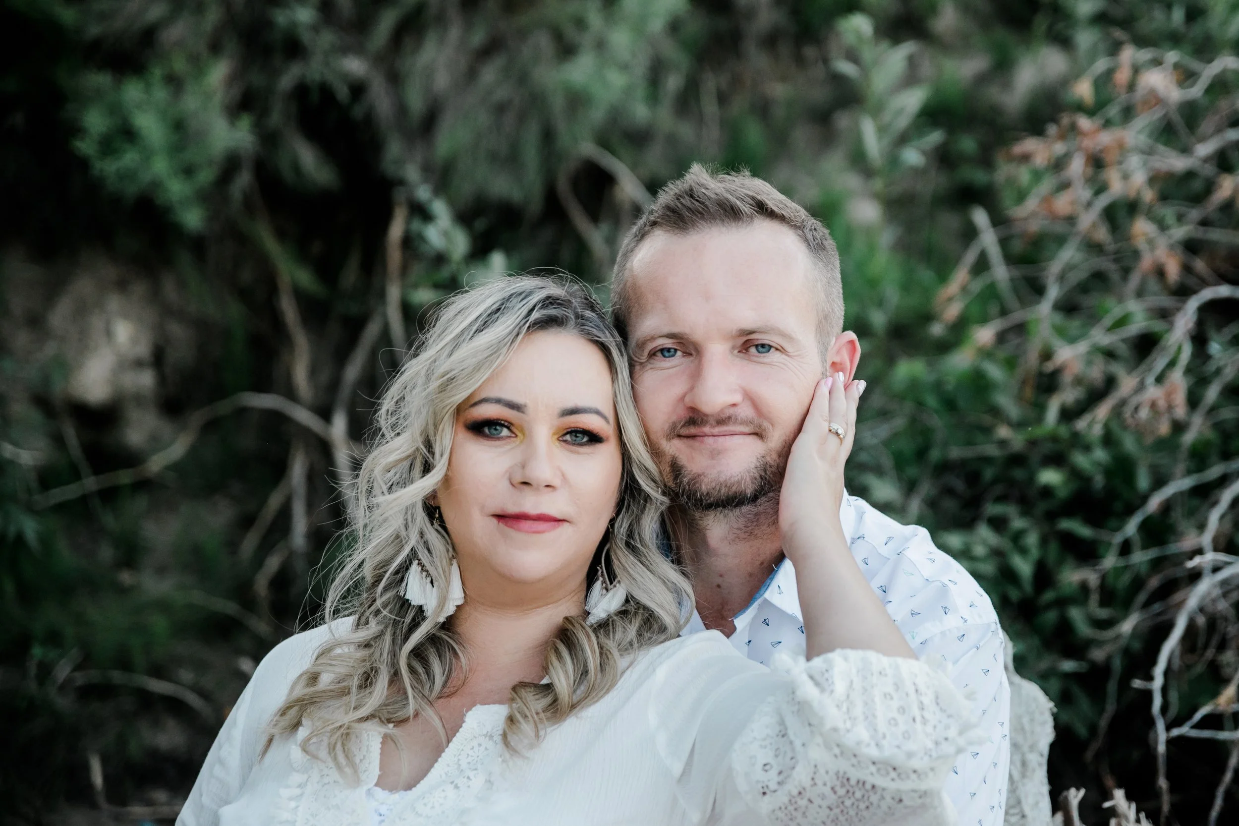A couples photo outdoors with a woman and man close together, the woman touching the man's face, with greenery in the background.