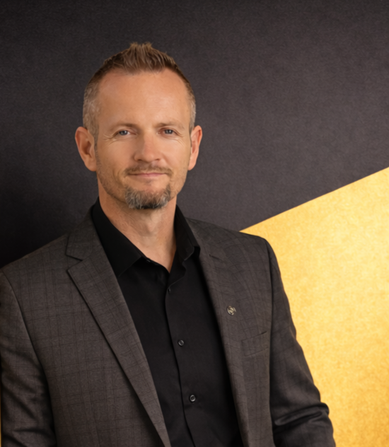Portrait of a middle-aged man with light skin, short light brown hair, a goatee, wearing a dark blazer and black shirt, standing in front of a dark background with a yellow geometric shape.