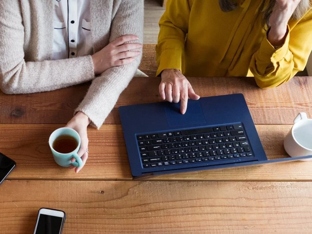 Two women sitting at a wooden table, one holding a coffee mug and the other pointing at a laptop.