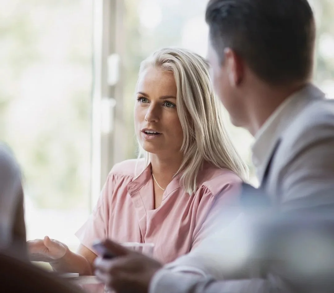 Two people are having a conversation indoors near a window. The woman on the left is blonde, wearing a pink top, and the man on the right has dark hair, wearing a white shirt, and is holding a phone.