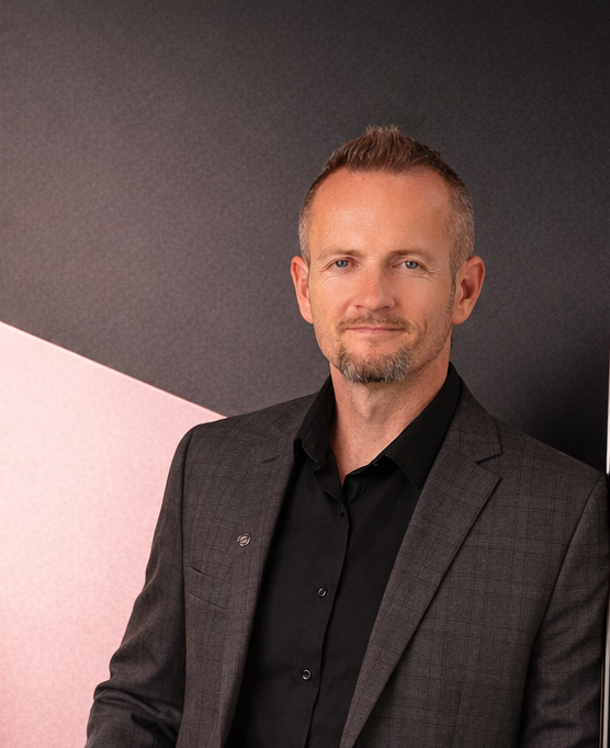 A man with short light brown hair, a light beard, wearing a dark suit jacket and black shirt, posing against a background with black and pink diagonal shapes.