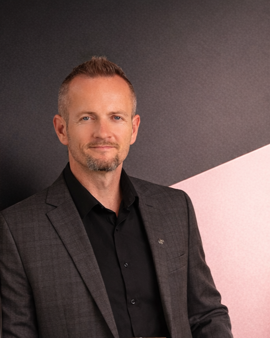 A man with short hair and a goatee, wearing a black shirt and a dark gray blazer, standing against a background with black and pink geometric shapes.