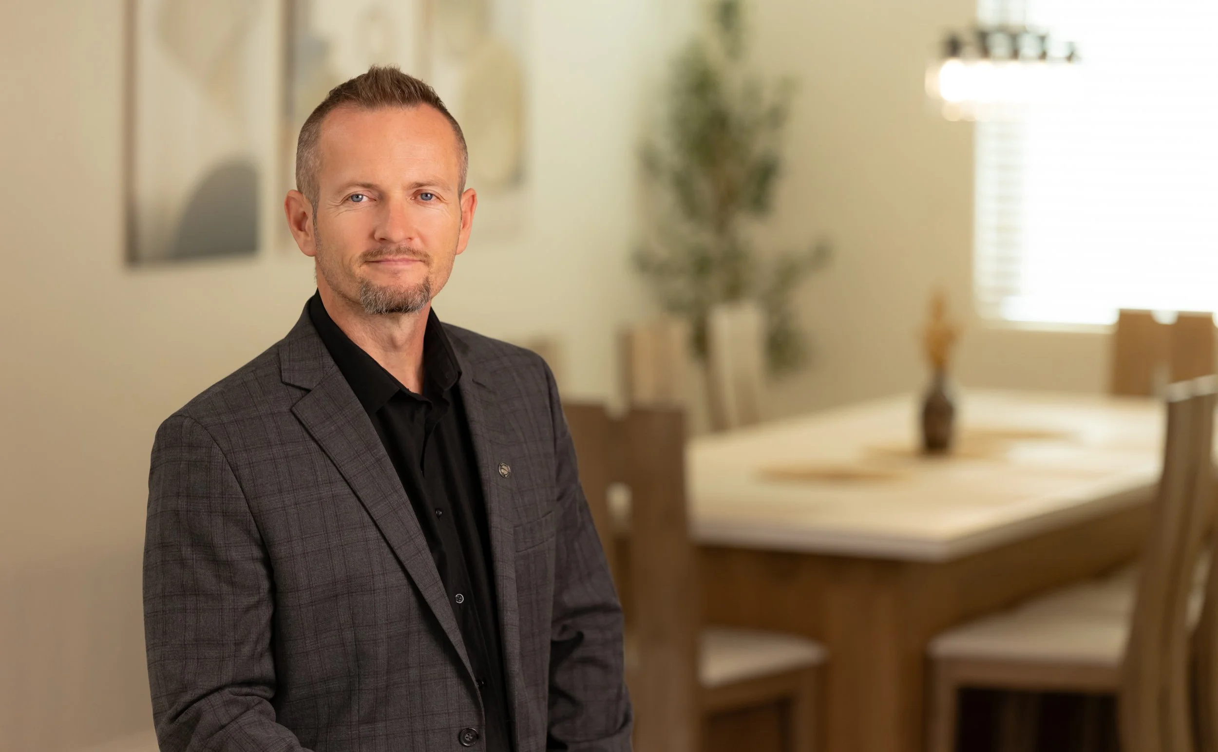 A middle-aged man in a dark checkered suit jacket and black shirt standing in a dining room with a wooden table and chairs, a vase with flowers, and window with blinds in the background.