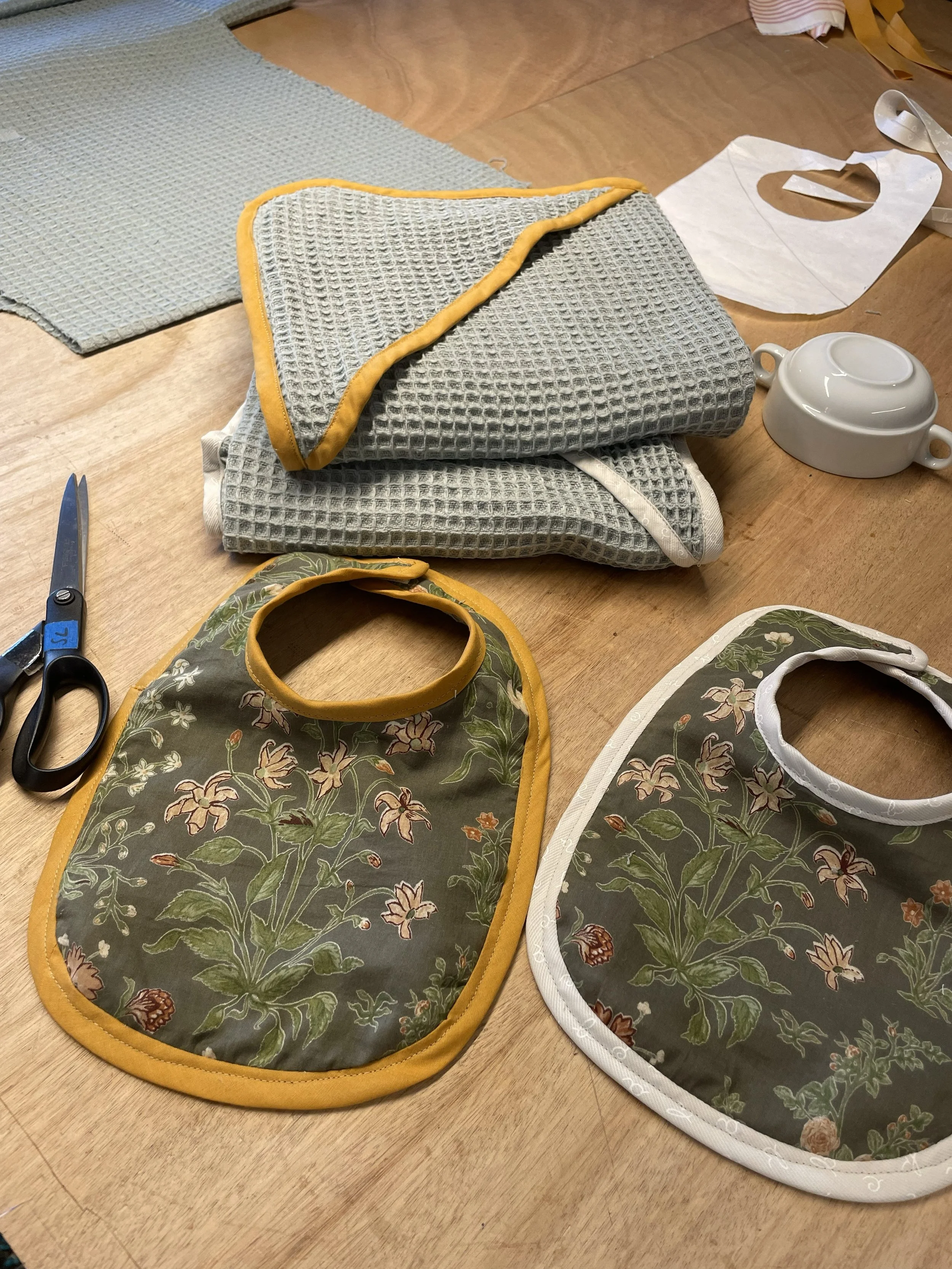 Différents linges de table et bavoirs pour bébé sur une table en bois, avec des ciseaux, une tasse en céramique et du papier en arrière-plan.