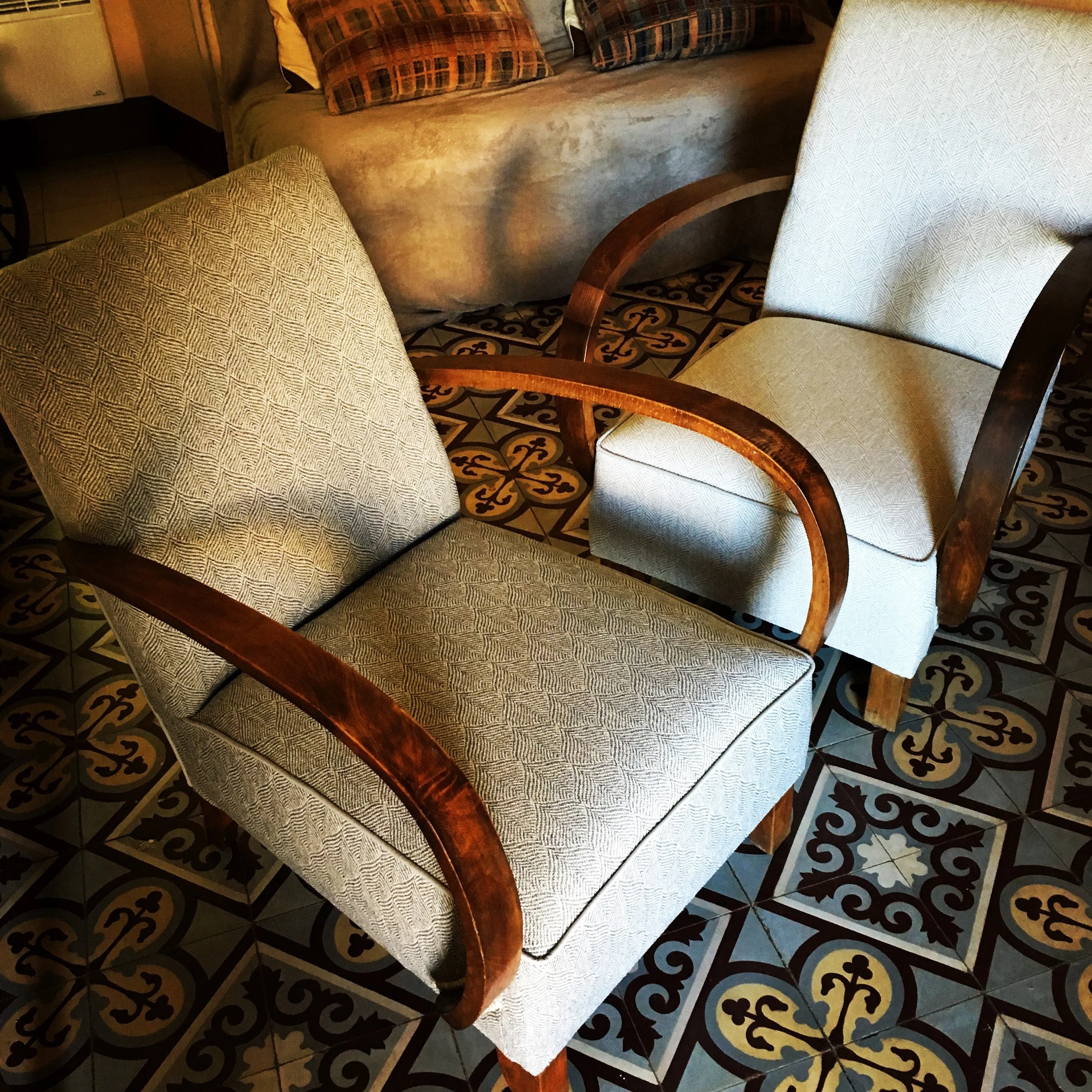 Deux fauteuils en bois avec coussins et garniture en tissu clair, placés sur un sol en carreaux décoratifs.