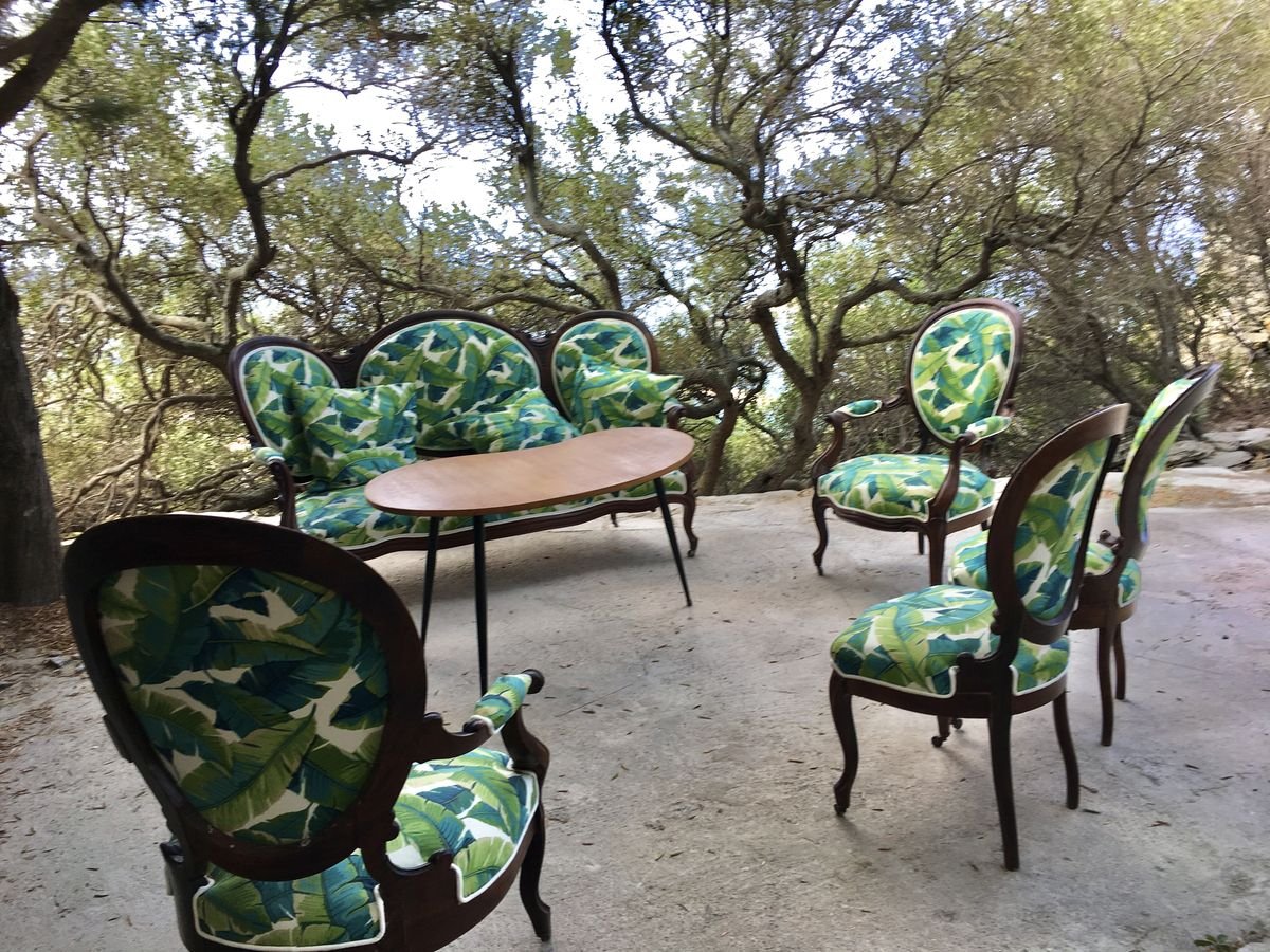Assortiment de meubles en bois avec tissu à motifs de feuilles vertes, comprenant un sofa, six chaises et une table, dans un environnement naturel sous des arbres.