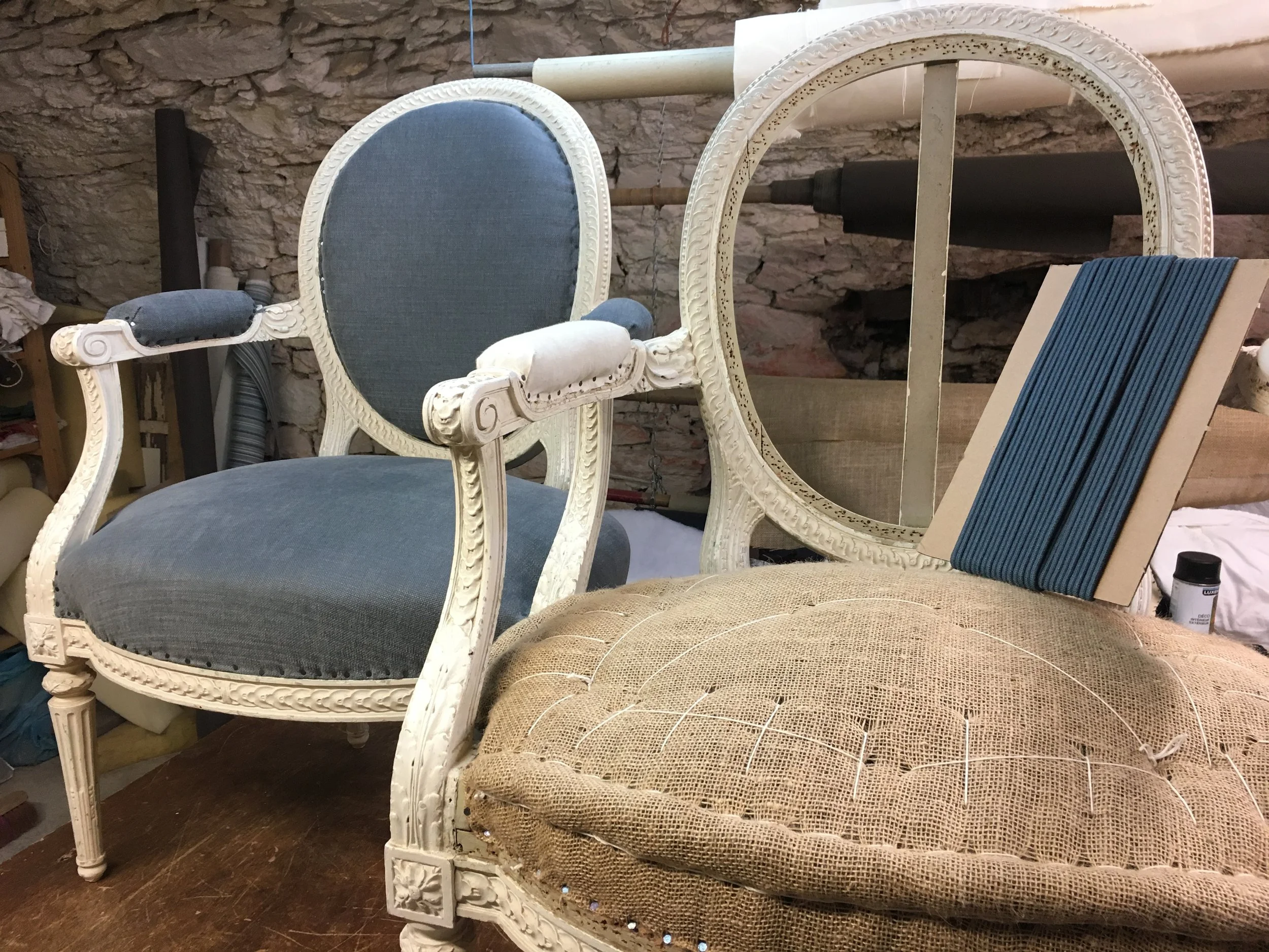 Deux fauteuils anciens en bois blanc, un avec un tissu bleu et l'autre avec un coussin en toile de jute, dans un atelier de restauration.