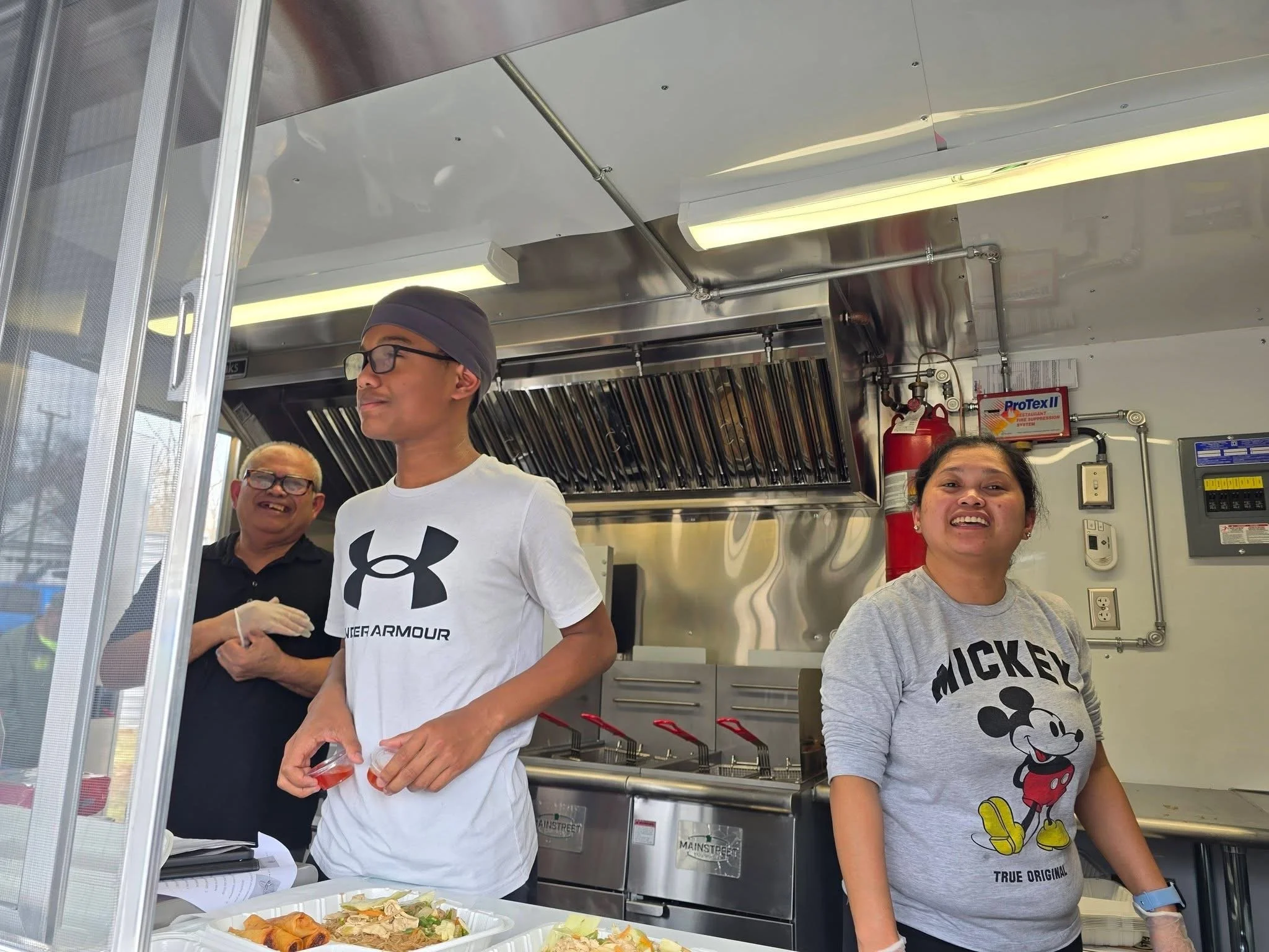 Three people inside a food truck or kitchen, two women and one man, all smiling and standing behind a counter with food items.