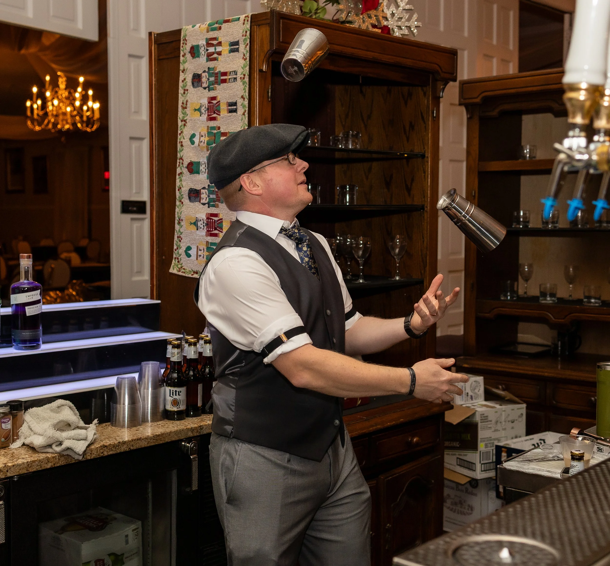 Bartender juggling three metal shakers in a bar setting with wooden shelves and bottles in the background.
