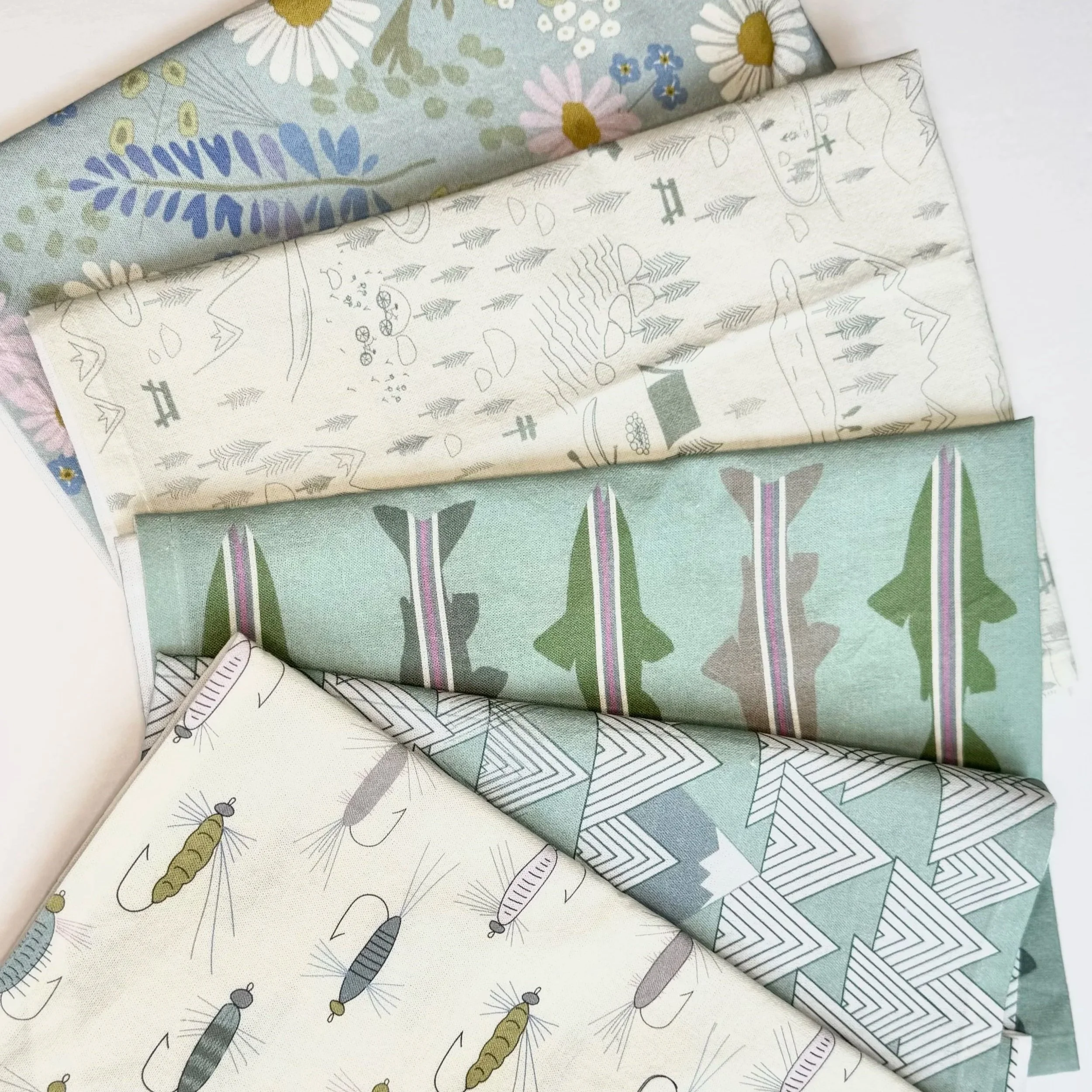 Five floded fabric tea towels with various nature-themed patterns, including flowers, mountains, and, rainbow trout, and fishing flies, arranged on a white surface.