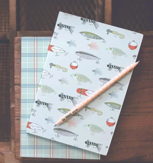 A notebook with a fishing-themed cover featuring fishing lures and fishing bobbers, topped with a pink pen, on a wooden surface.