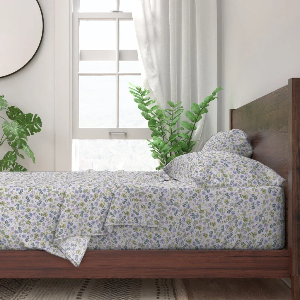 A bedroom with a wooden bed frame, floral bedding, a large green plant, a window with white curtains, and a round mirror on the wall.