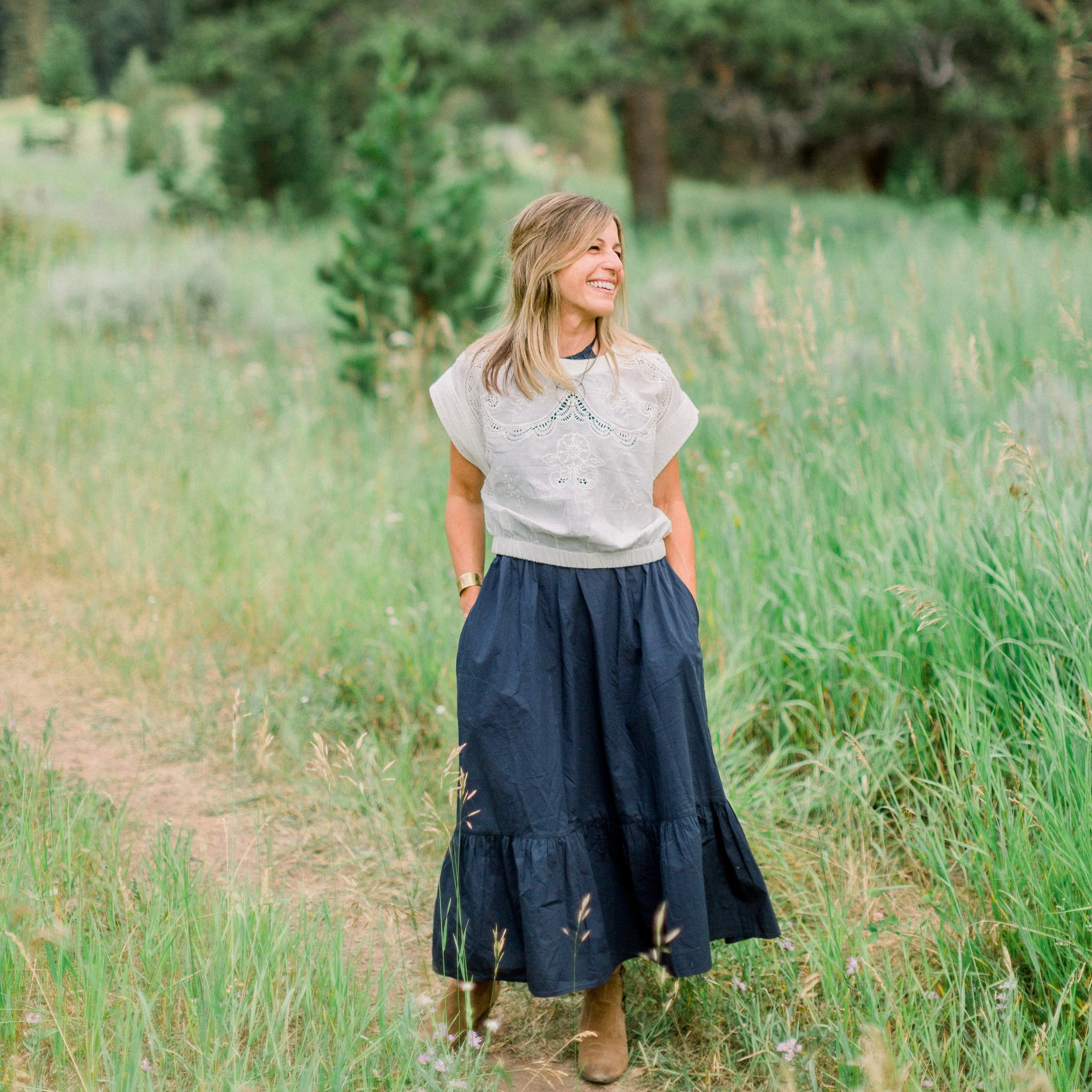 Woman in a white embroidered top and long navy skirt standing on a grassy path in a green field, smiling and looking to the side.