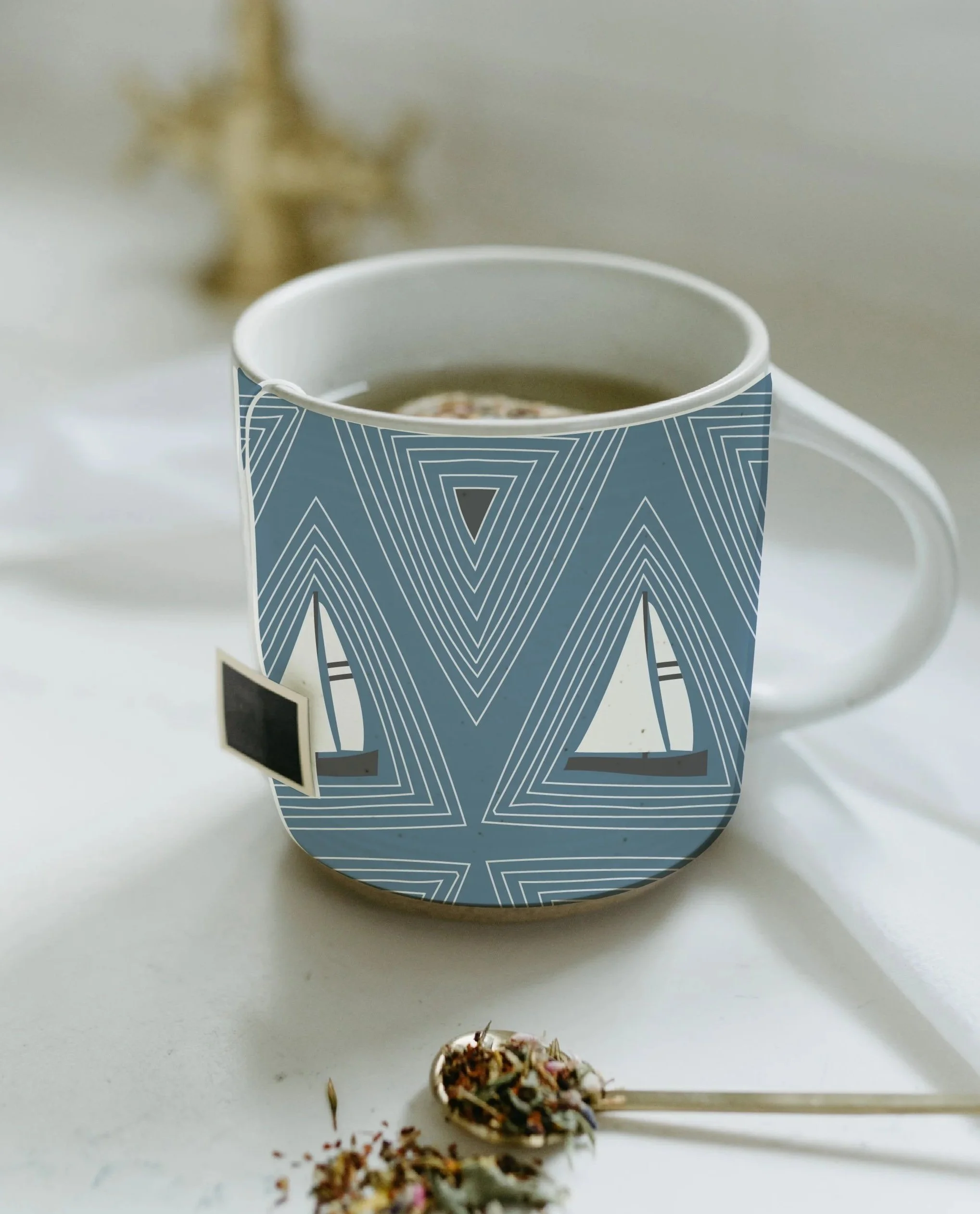 A ceramic mug with a geometric blue and white sailboat pattern, filled with a hot beverage, placed on a white surface with a tea infuser with loose tea and herbs nearby.