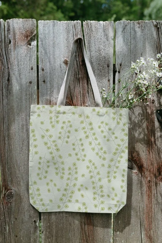 A reusable tote bag with green leaf patterns hanging on a wooden fence, with some small white flowers visible on the right.