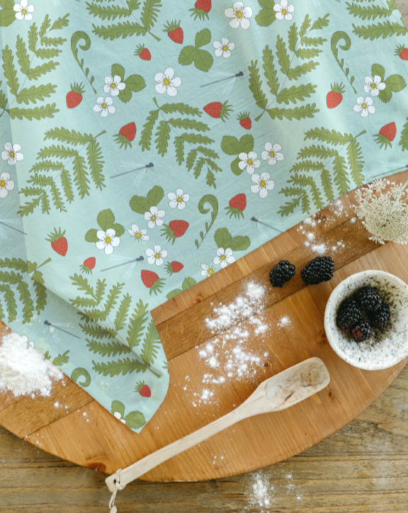 A wooden round cutting board with a blue cloth featuring strawberries, leaves, and white flowers and dragonflies, some blackberries in a small bowl, a wooden spoon, and scattered flour on the board and table.