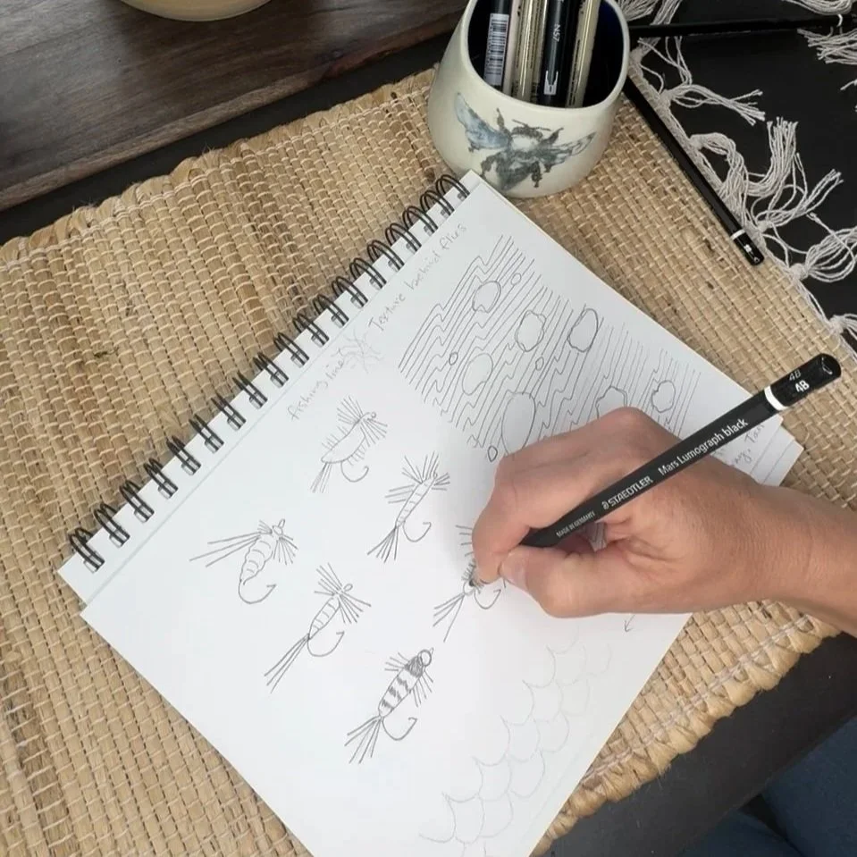 A person sketches various fly fishing lure illustrations and pattern designs in a notebook on a woven mat, with pens and markers nearby.