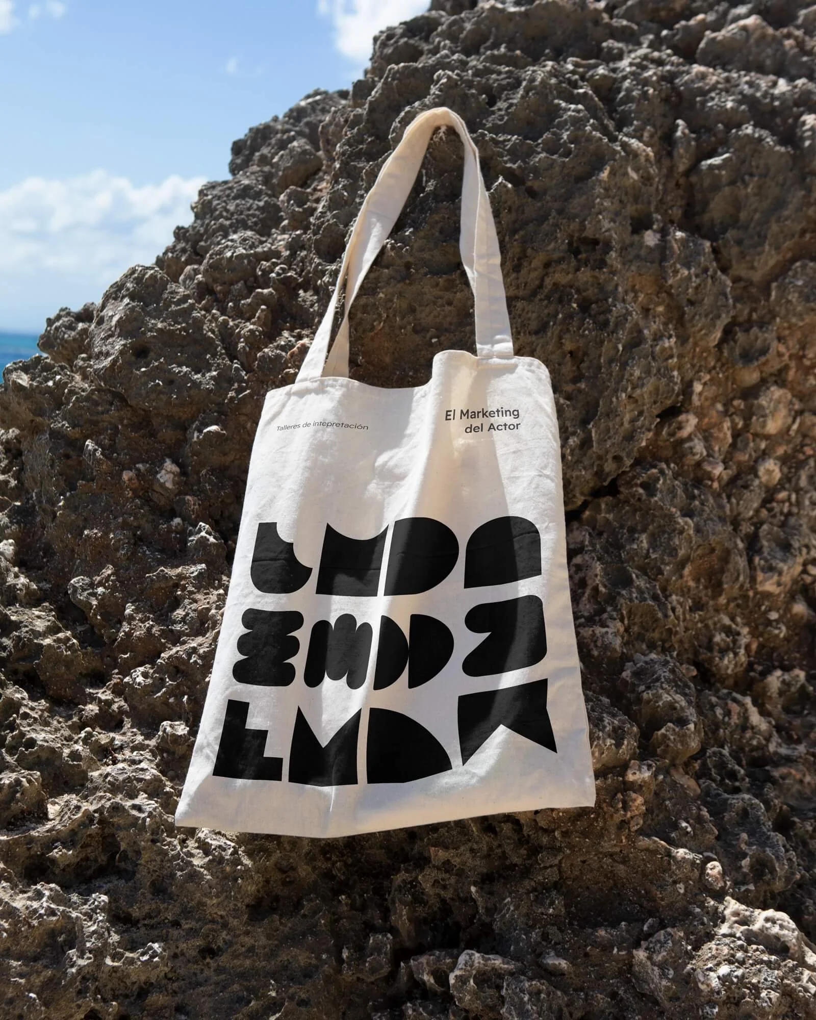 Bolsa de tela blanca colgada de una roca , con texto en la bolsa que dice 'El Marketing del Actor' y 'Talleres de interpretación', además de letras grandes y negras en forma de bloques en las que aparecen las siglas "EMDA"