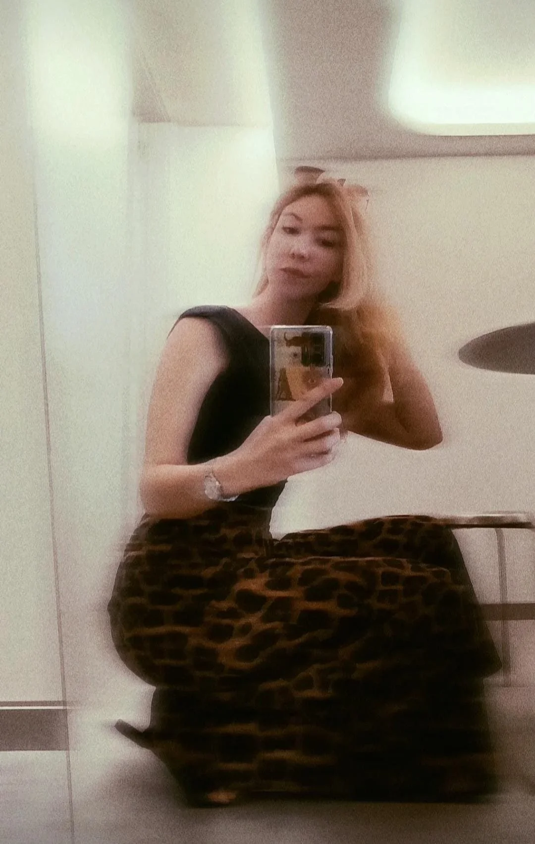 Mujer tomando una selfie en un espejo, usando un vestido negro y pantalones de leopardo, con cabello rubio.