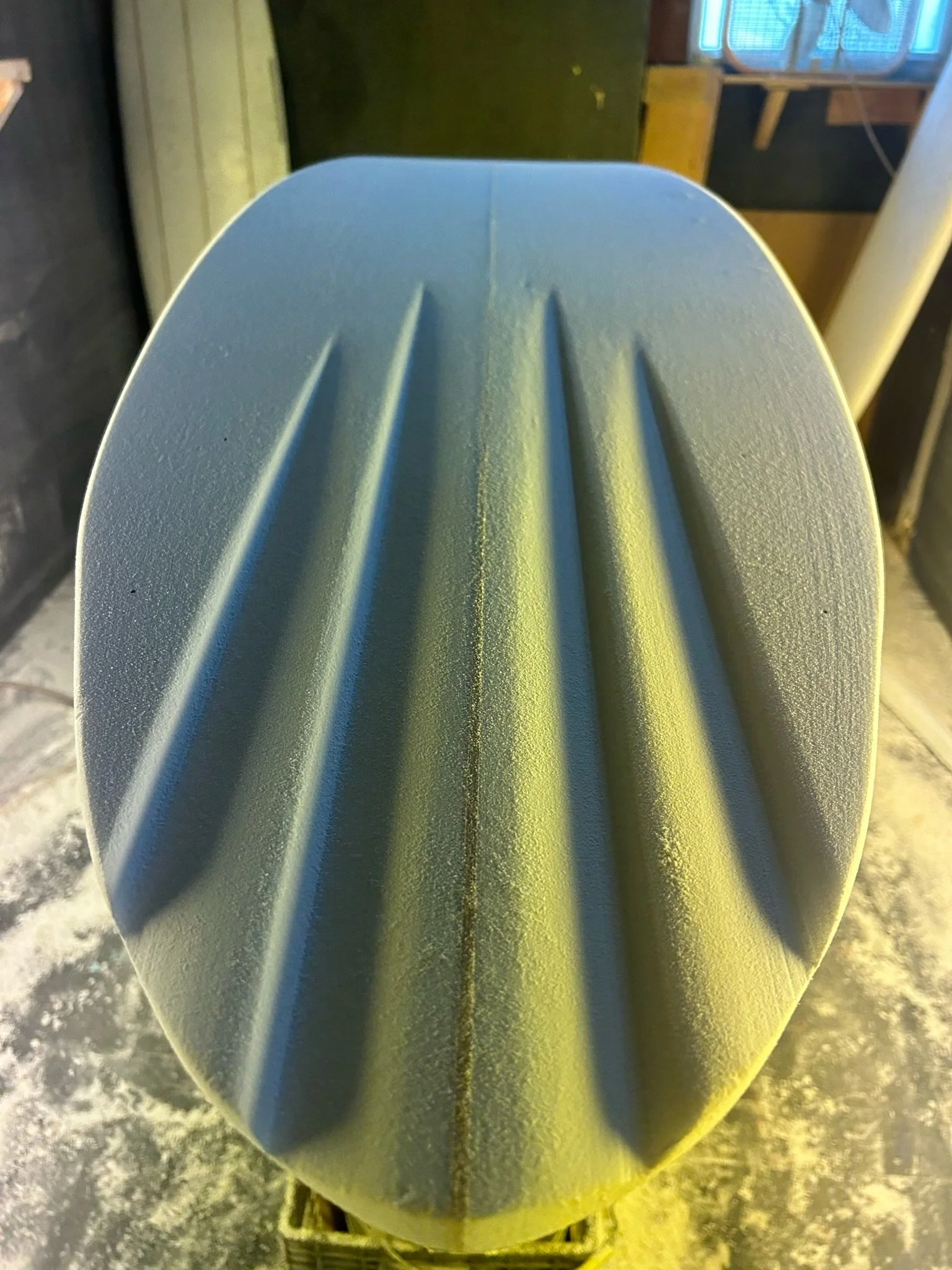 Close-up of a channel bottom surfboard, positioned vertically on a workbench in a workshop with tools and equipment in the background.