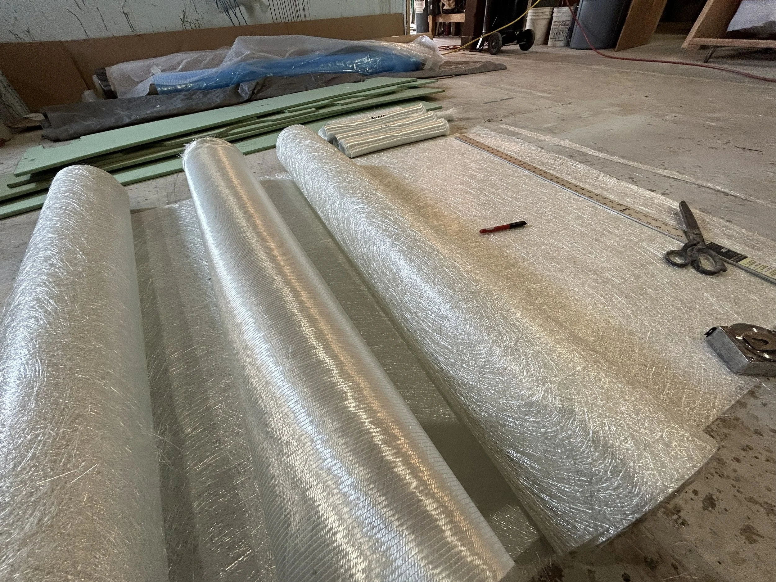 Construction materials including rolls of reflective insulation, green foam boards, and a measuring tape laid out on a wooden floor in a construction site. Fiberglass chopped strand matt and woven cloth.