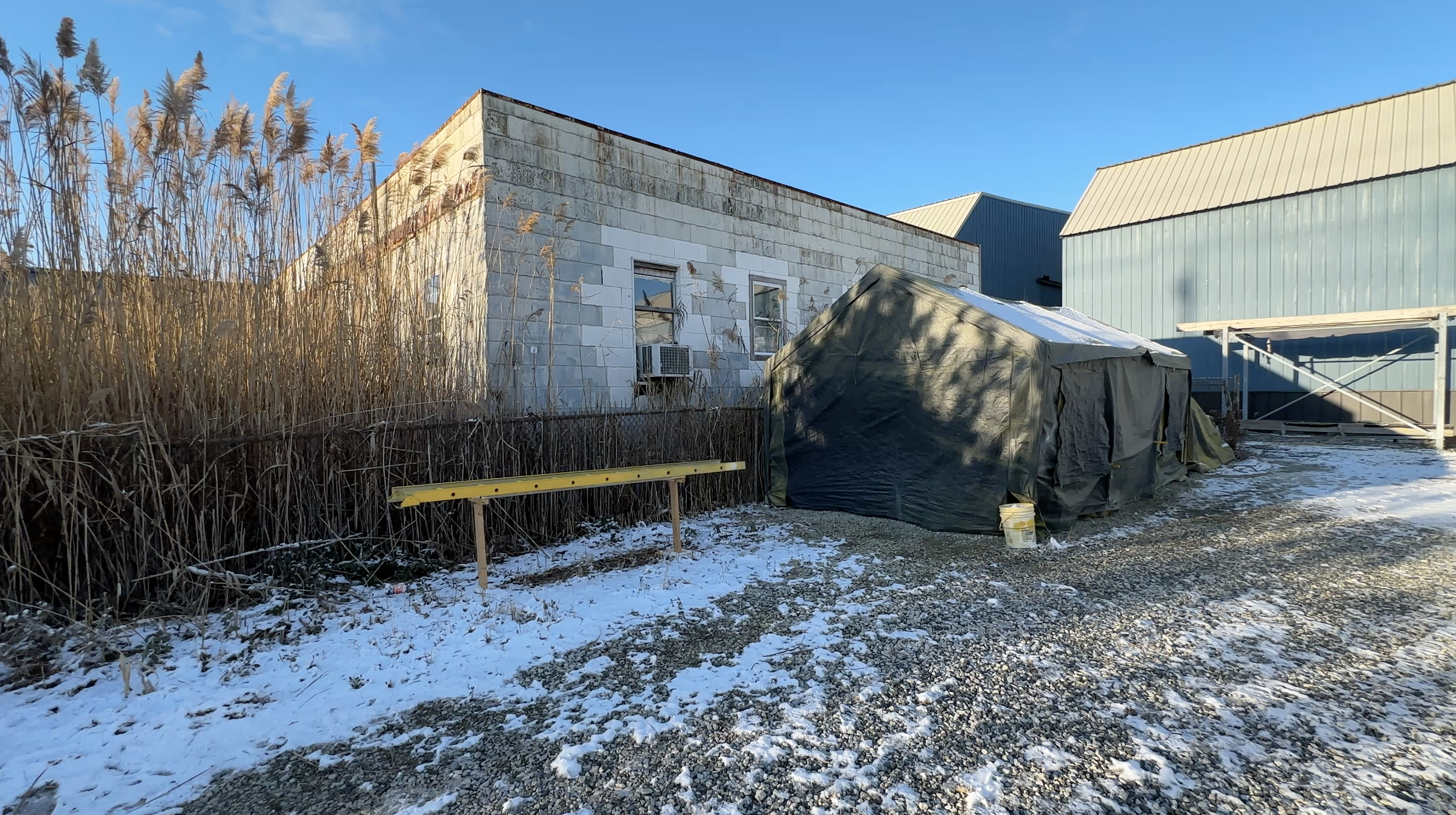 An under-construction building with masonry walls and a green tarp covering construction materials outdoors, with snow on the ground and tall dry grass to the side, under a clear blue sky.