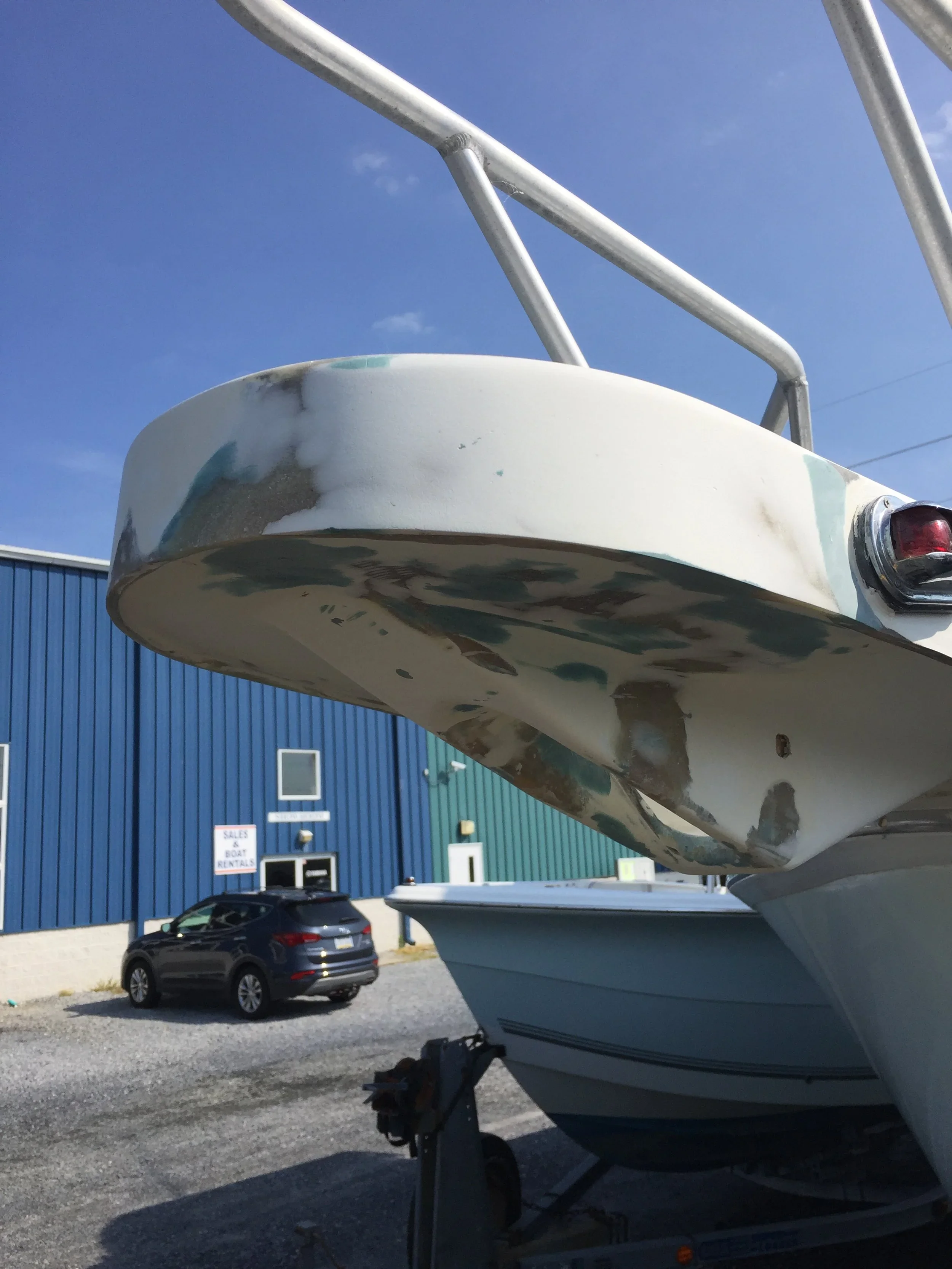 A boat on a trailer at a boat rental or sales lot with a blue building in the background and a parked car in the foreground, under a blue sky.