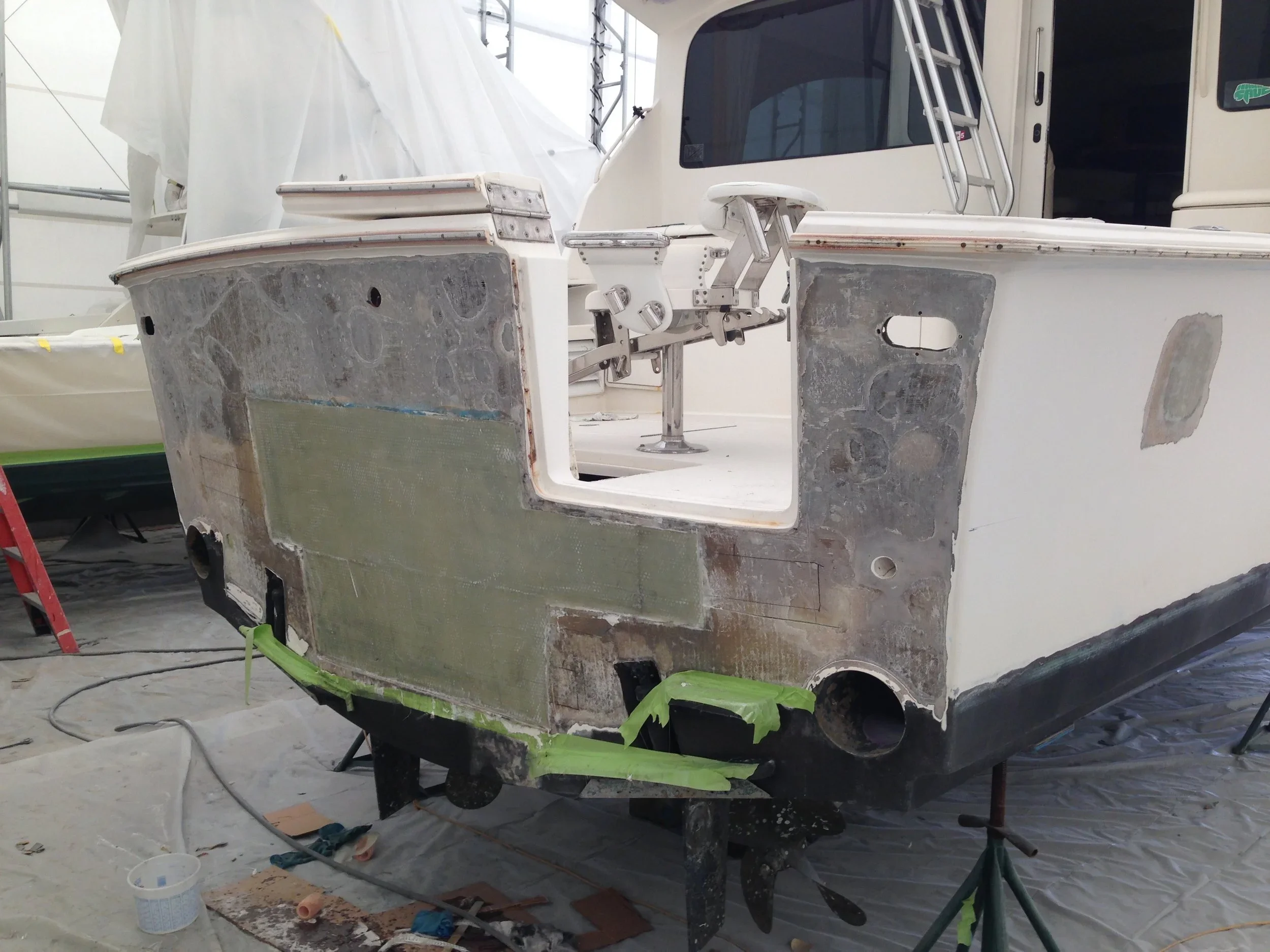 The image shows a boat undergoing restoration or repair inside a workshop, with visible sanding and patchwork on its hull, surrounded by tools and materials.