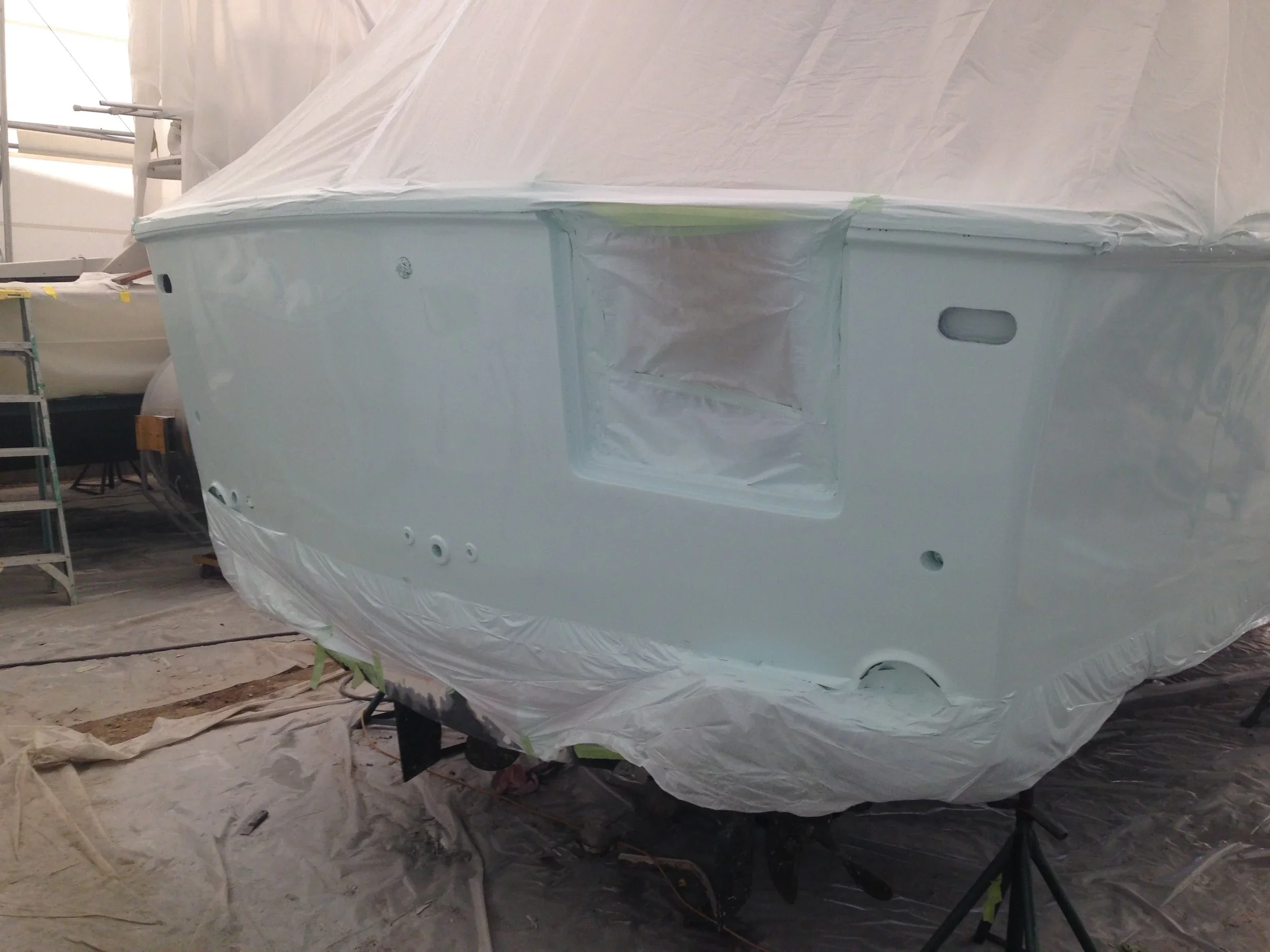 A boat being painted or repaired inside a workshop, covered with masking paper and tape, with some scaffolding and tools visible in the background.